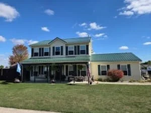 Two-story home with green standing seam metal roof, dormers, and matching porch roof over pale yellow siding