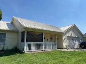 Single-story home with light gray standing-seam metal roof, white porch railings, and beige vinyl siding over a green lawn